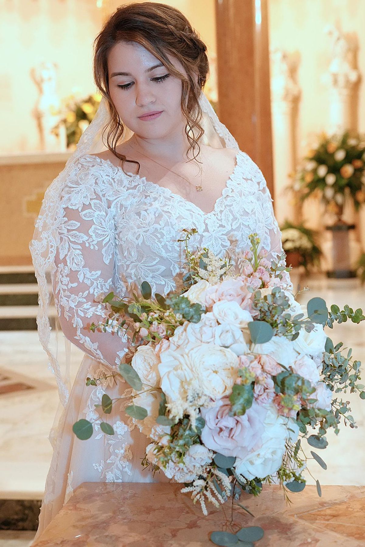 A bride in a lace gown looks down at her blush and ivory bouquet