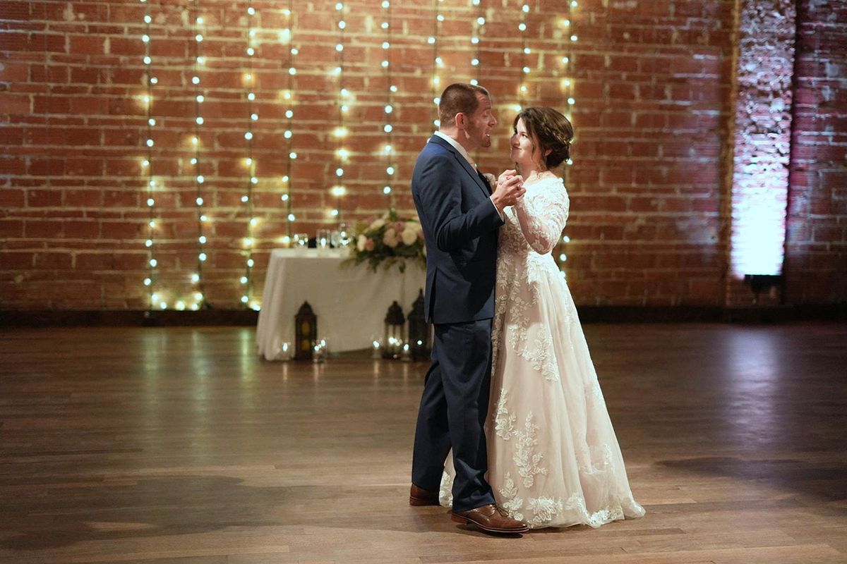 Newlyweds share their first dance beneath strings of warm lights and a brick wall