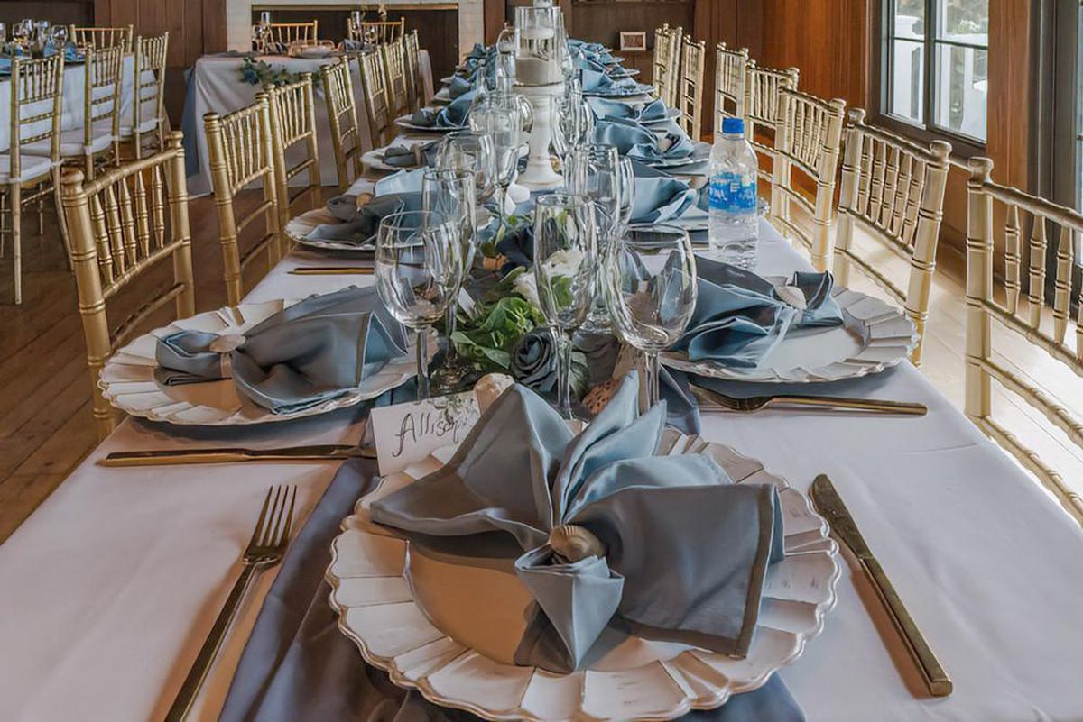 A long reception table set with dusty blue napkins, gold flatware and glassware