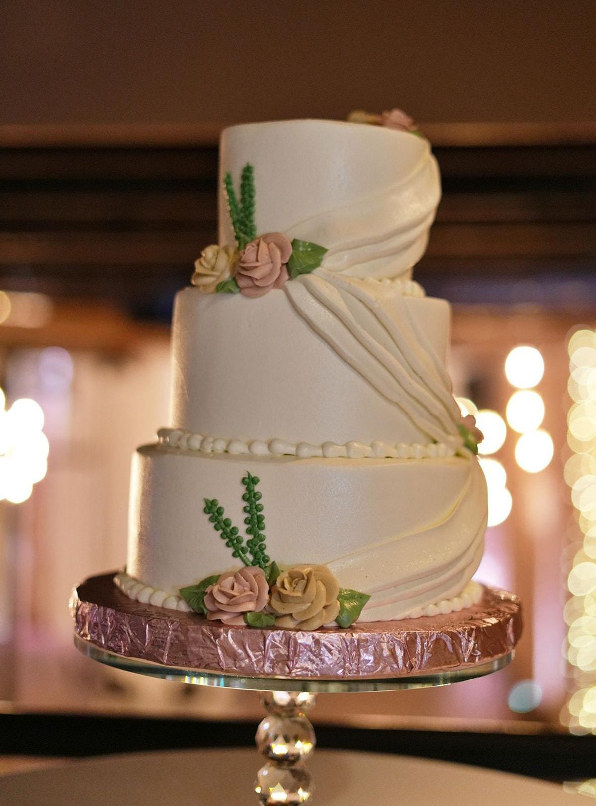 A draped three-tier wedding cake with blush sugar roses against glowing reception lights